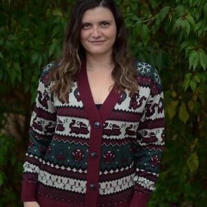 May include: A burgundy, green, and white patterned cardigan sweater with a V-neck and button closure. The sweater features a festive design with snowflakes and other holiday-themed motifs. The model is wearing blue jeans.