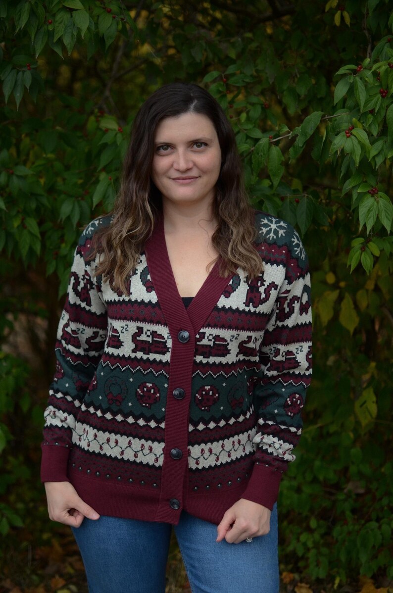 May include: A burgundy, green, and white patterned cardigan sweater with a V-neck and button closure. The sweater features a festive design with snowflakes and other holiday-themed motifs. The model is wearing blue jeans.