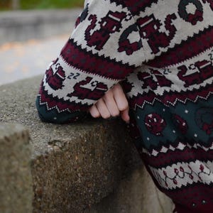 May include: A festive Christmas sweater in green, burgundy, and cream. The sweater features pixelated holiday designs, including snowflakes, reindeer, and wreaths. The sweater has a crew neck and long sleeves, perfect for the holiday season.