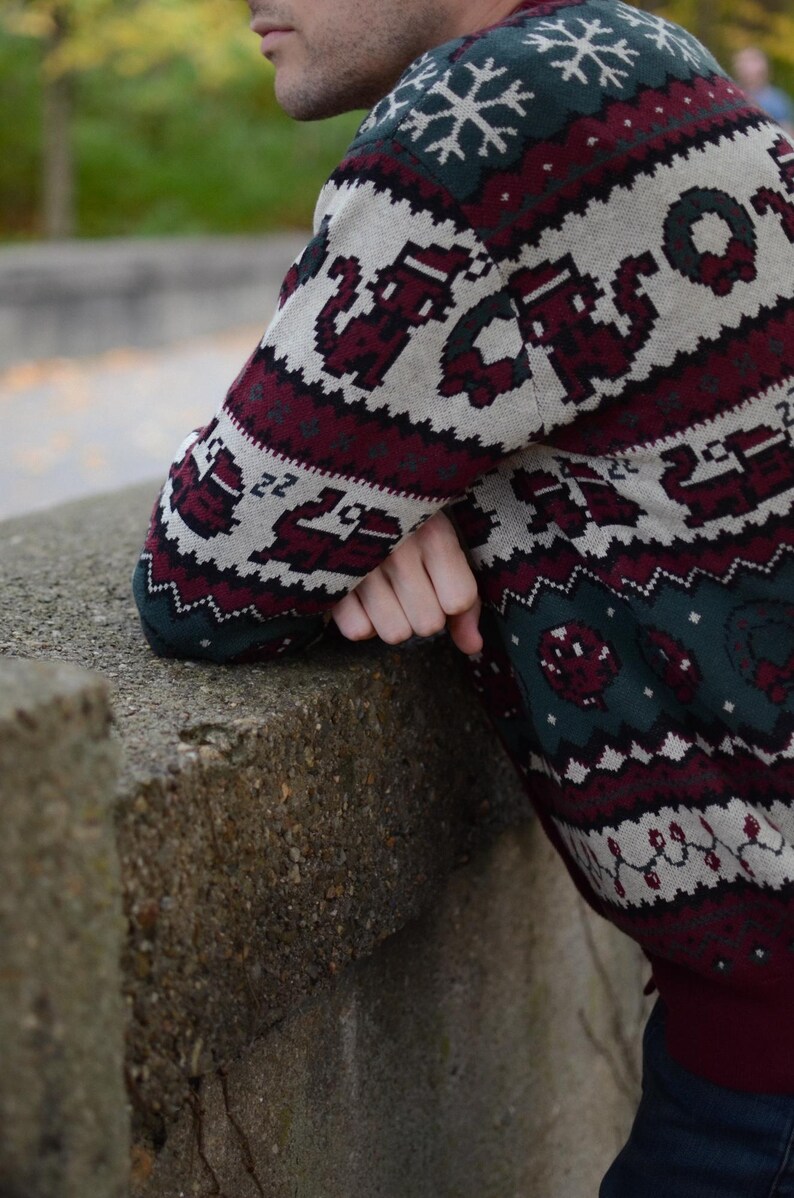 May include: A festive Christmas sweater in green, burgundy, and cream. The sweater features pixelated holiday designs, including snowflakes, reindeer, and wreaths. The sweater has a crew neck and long sleeves, perfect for the holiday season.