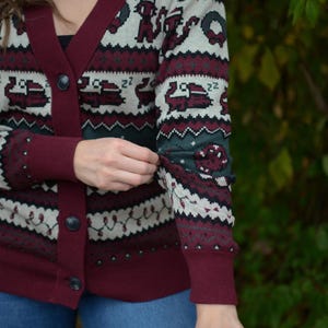 May include: Burgundy cardigan sweater with a festive pattern. The sweater features a V-neck, button closure, and ribbed cuffs. The pattern includes snowflakes, Christmas trees, and other holiday-themed designs in white, green, and burgundy.