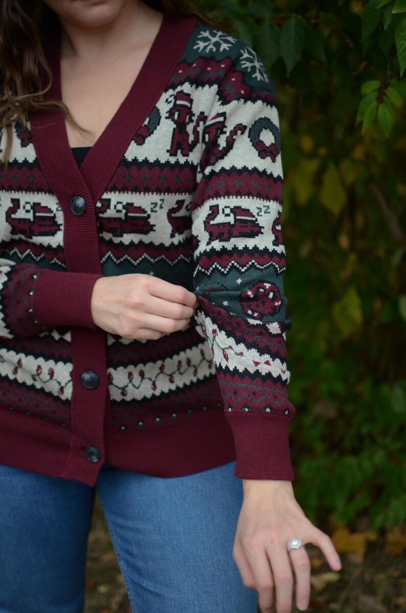 May include: Burgundy cardigan sweater with a festive pattern. The sweater features a V-neck, button closure, and ribbed cuffs. The pattern includes snowflakes, Christmas trees, and other holiday-themed designs in white, green, and burgundy.