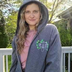 Op de afbeelding: Een grijze hoodie met rits met een groen ontwerp van een videogamecontroller op de voorkant. De controller is omringd door groene bladeren.