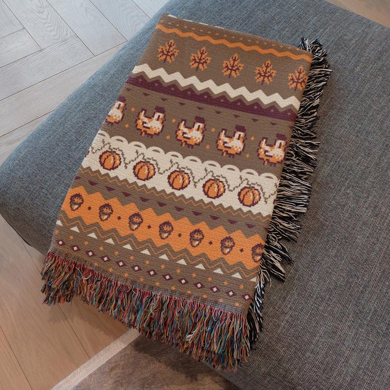 May include: A brown throw blanket with a fall-themed pattern. The design includes pumpkins, acorns, and leaves in shades of orange, white, and burgundy. The blanket has a fringed edge with multiple colors. The blanket is folded on a gray surface.
