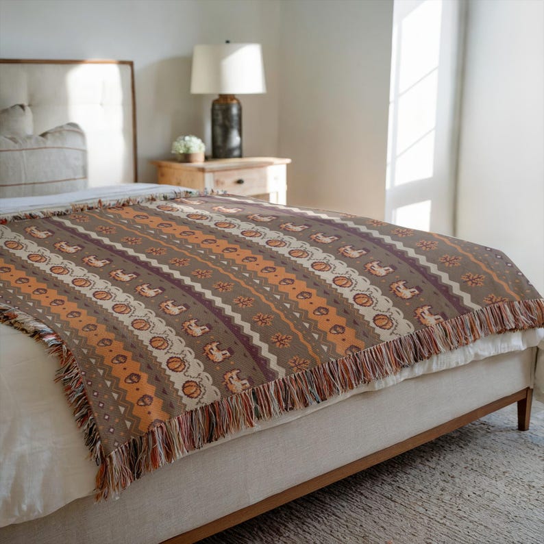 May include: A brown and orange patterned throw blanket with fringe. The blanket features a repeating pattern of stylized pumpkins, flowers, and geometric shapes in shades of brown, orange, and cream. The blanket is draped over a bed.
