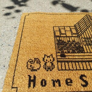 Puede incluir: Una alfombra de puerta rectangular hecha de fibra de coco marr&oacute;n natural. La alfombra presenta un gr&aacute;fico negro de una casa, dos personajes de dibujos animados pixelados y las palabras "Home Sweet". La alfombra est&aacute; dise&ntilde;ada para uso en exteriores.