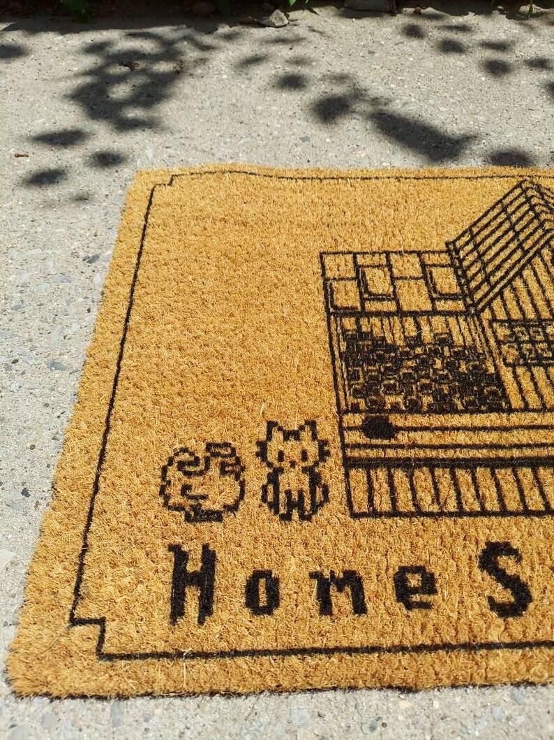 Puede incluir: Una alfombra de puerta rectangular hecha de fibra de coco marr&oacute;n natural. La alfombra presenta un gr&aacute;fico negro de una casa, dos personajes de dibujos animados pixelados y las palabras "Home Sweet". La alfombra est&aacute; dise&ntilde;ada para uso en exteriores.