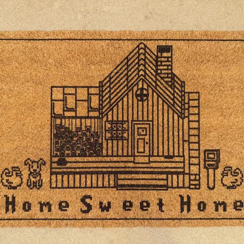 May include: A rectangular doormat with a house illustration and the words "Home Sweet Home" in black. The house design includes a chimney, windows, and a door. Two small animal figures are on the left side of the mat.