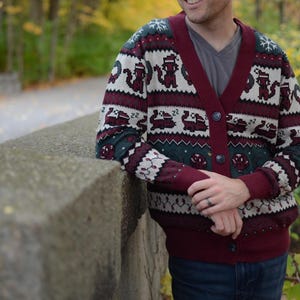 May include: A burgundy and cream-colored cardigan sweater with a repeating pattern of cartoon animals and snowflakes. The sweater has a V-neck and button closure. The person is wearing a gray t-shirt and blue jeans.