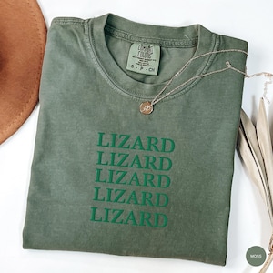 Puede incluir: Una camiseta verde musgo con la palabra "LIZARD" impresa cinco veces en verde. Un collar de oro con un colgante está drapeado sobre la camiseta. La camiseta está doblada sobre una superficie blanca.