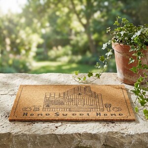 May include: A rectangular doormat made of natural fibers. The mat features a detailed black line drawing of a house with the words "Home Sweet Home" beneath it. A potted plant sits to the right of the mat.