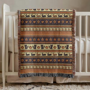 May include: A Halloween-themed throw blanket with a repeating pattern of black cats, pumpkins, and leaves in shades of orange, brown, and cream. The blanket has a fringed edge and is draped over a white crib.