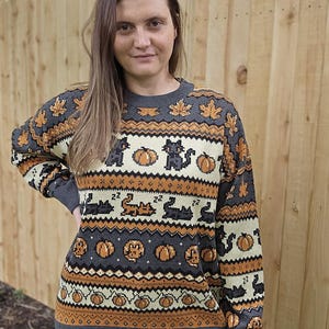 Puede incluir: Un su&eacute;ter gris de Halloween con detalles en naranja y crema. El su&eacute;ter presenta un patr&oacute;n de calabazas, gatos negros y hojas de arce. Los pu&ntilde;os y el dobladillo son de color gris liso.