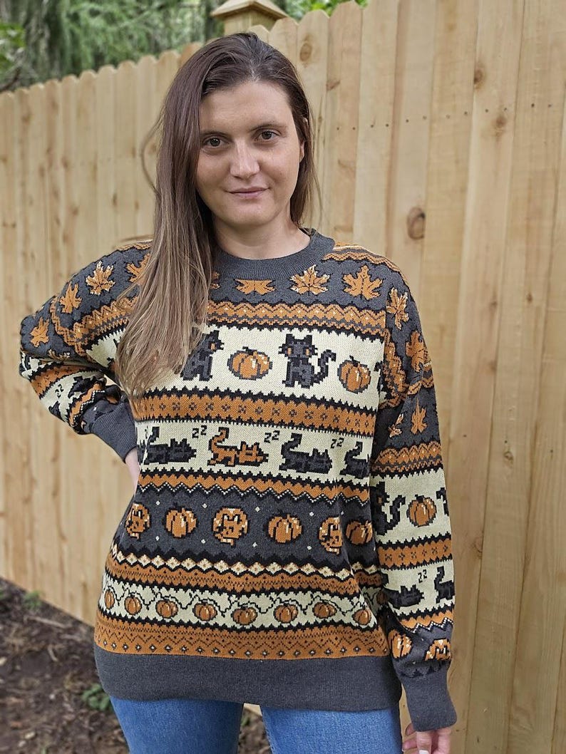 Puede incluir: Un su&eacute;ter gris de Halloween con detalles en naranja y crema. El su&eacute;ter presenta un patr&oacute;n de calabazas, gatos negros y hojas de arce. Los pu&ntilde;os y el dobladillo son de color gris liso.