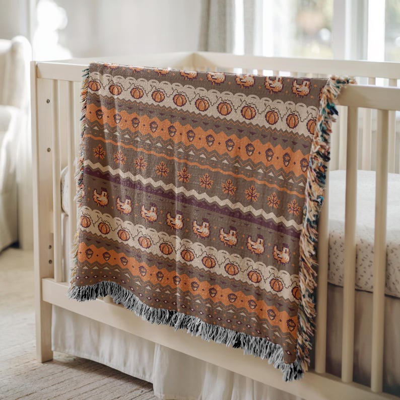May include: A woven throw blanket with a fall-themed pattern. The blanket features repeating designs of pumpkins, acorns, and other autumn motifs in shades of brown, orange, and cream. The blanket is draped over a white crib.