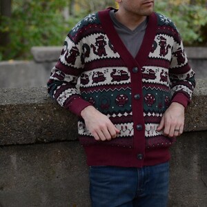 May include: Burgundy cardigan sweater with a festive pattern featuring repeating figures, snowflakes, and geometric shapes. The sweater has a V-neck, button closure, and dark green and white accents. The person is wearing blue jeans.