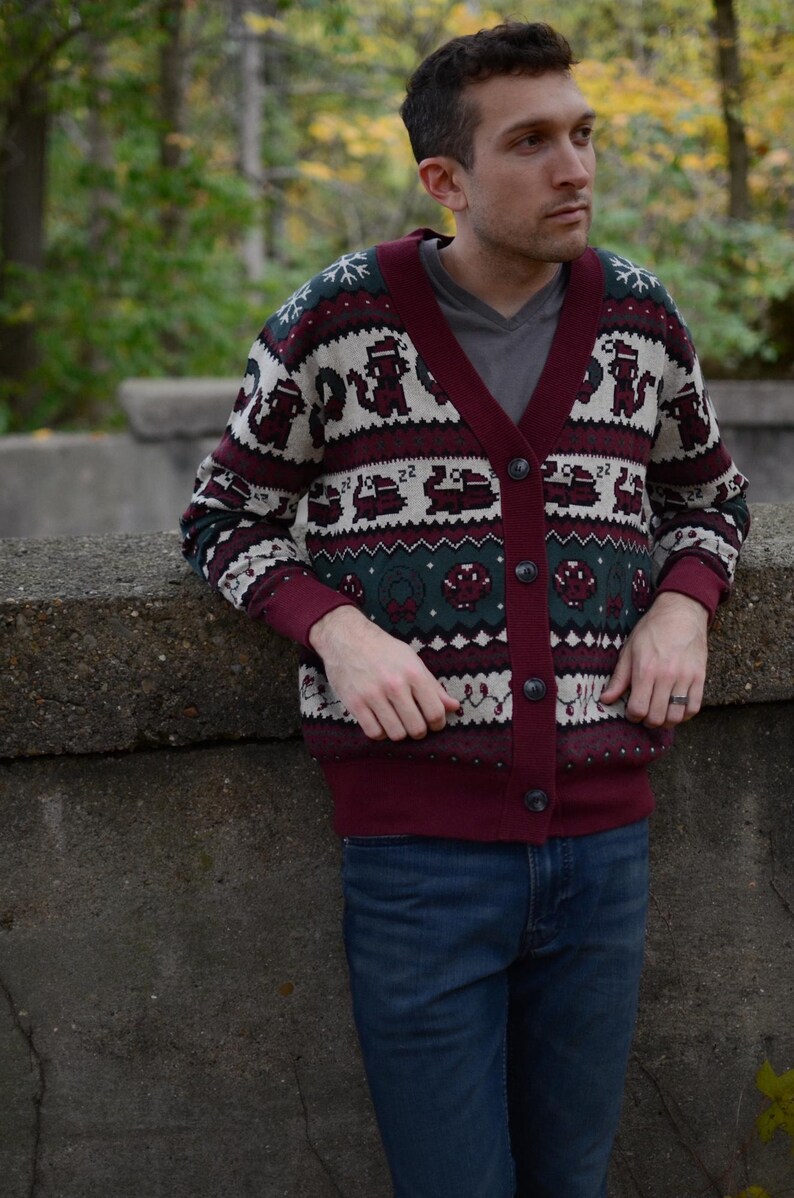 May include: Burgundy cardigan sweater with a festive pattern featuring repeating figures, snowflakes, and geometric shapes. The sweater has a V-neck, button closure, and dark green and white accents. The person is wearing blue jeans.