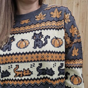 Puede incluir: Su&eacute;ter gris con un patr&oacute;n repetido de calabazas, gatos negros y hojas de oto&ntilde;o en colores naranja y crema. El su&eacute;ter tiene cuello redondo y mangas largas, perfecto para el oto&ntilde;o.