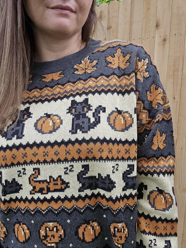 Puede incluir: Su&eacute;ter gris con un patr&oacute;n repetido de calabazas, gatos negros y hojas de oto&ntilde;o en colores naranja y crema. El su&eacute;ter tiene cuello redondo y mangas largas, perfecto para el oto&ntilde;o.