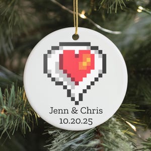 Peut inclure: Ornement de Noël rond en céramique blanche avec un motif de cœur pixélisé rouge, orange et noir. Les noms "Jenn & Chris" et la date "10.20.25" sont imprimés en noir. Une ficelle dorée est attachée pour suspendre.