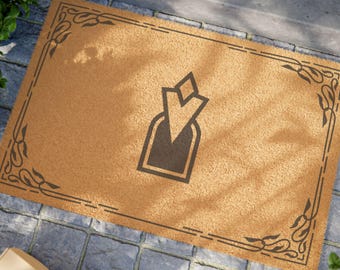 May include: A brown coir doormat with a black and white design of a stylized arrow in a square frame. The doormat has a decorative border with a floral pattern.
