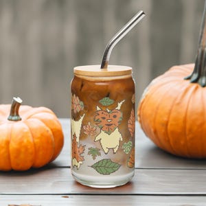 May include: A glass can-shaped tumbler with a wooden lid and a metal straw, filled with a layered coffee drink. The glass is decorated with a pixelated cartoon character and fall leaves. Two orange pumpkins sit nearby, suggesting an autumn theme.