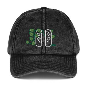 May include: A black vintage washed denim baseball cap with an embroidered design of two video game controllers with green vines growing around them.