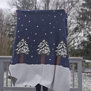 May include: A navy blue blanket with a winter scene design. The blanket features white pixelated stars and three Christmas trees with brown trunks and white and green pixelated branches. A gray border is at the bottom.
