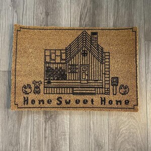 May include: Rectangular doormat with a brown coir surface and a black outline of a house, dog, and mailbox. The words "Home Sweet Home" are printed in black across the bottom of the mat.