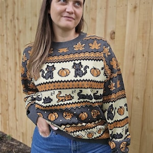 Puede incluir: Un su&eacute;ter gris con un patr&oacute;n de Halloween con gatos negros, calabazas y hojas de arce en colores naranja y crema. El su&eacute;ter tiene cuello redondo y mangas largas.