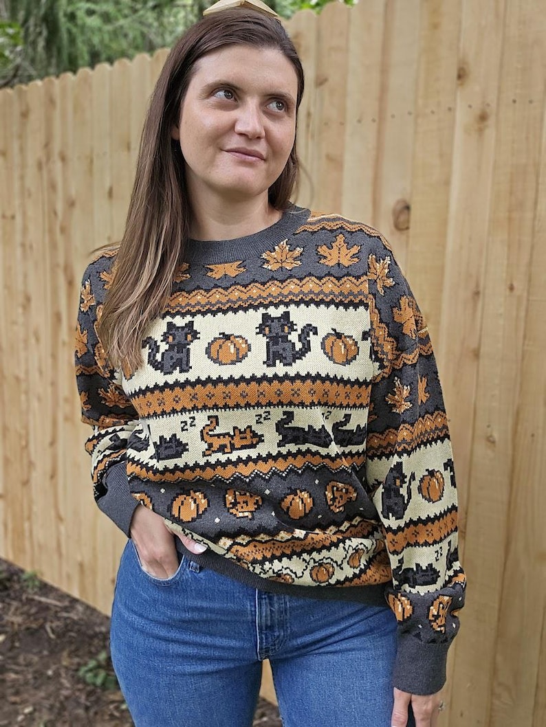 Puede incluir: Un su&eacute;ter gris con un patr&oacute;n de Halloween con gatos negros, calabazas y hojas de arce en colores naranja y crema. El su&eacute;ter tiene cuello redondo y mangas largas.