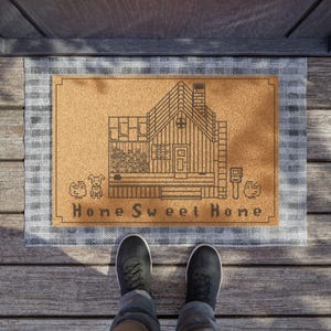 May include: A rectangular doormat with a brown center and a gray plaid border. The center features a line drawing of a house with the words "Home Sweet Home" below. A person is standing on the mat.