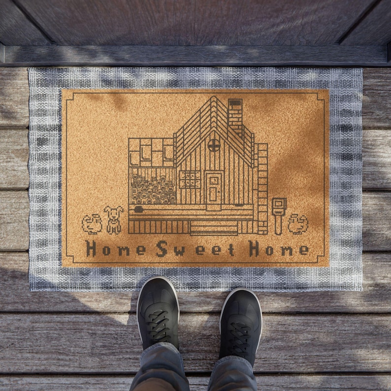 May include: A rectangular doormat with a brown center and a gray plaid border. The center features a line drawing of a house with the words "Home Sweet Home" below. A person is standing on the mat.