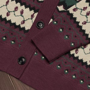 May include: A burgundy cardigan sweater with a patterned design in cream, green, and black. The sweater has a ribbed cuff and hem, and two black buttons. The pattern includes floral and geometric shapes.