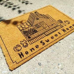 Puede incluir: Felpudo rectangular hecho de fibras naturales de coco. La alfombra presenta un dibujo lineal negro de una casa con las palabras "Home Sweet Home" en negrita, en negro. Los elementos decorativos incluyen un gato y un perro.