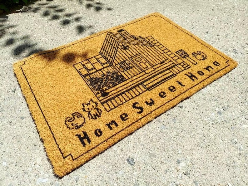 Puede incluir: Felpudo rectangular hecho de fibras naturales de coco. La alfombra presenta un dibujo lineal negro de una casa con las palabras "Home Sweet Home" en negrita, en negro. Los elementos decorativos incluyen un gato y un perro.