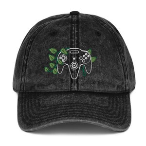 Puede incluir: Una gorra de béisbol de mezclilla vintage negra lavada con un controlador Nintendo 64 blanco bordado con enredaderas verdes.