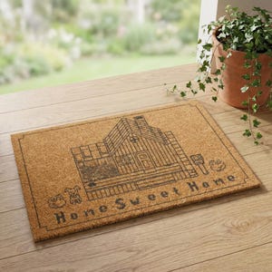 May include: Rectangular doormat made of natural coir fiber. The mat features a detailed black line drawing of a house with the words "Home Sweet Home" below the house. The mat is placed on a wooden surface.