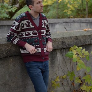 May include: A burgundy cardigan sweater with a festive pattern of reindeer, snowflakes, and other holiday motifs. The sweater has a button-up front and is paired with blue jeans. The background features a natural outdoor setting with trees and foliage.