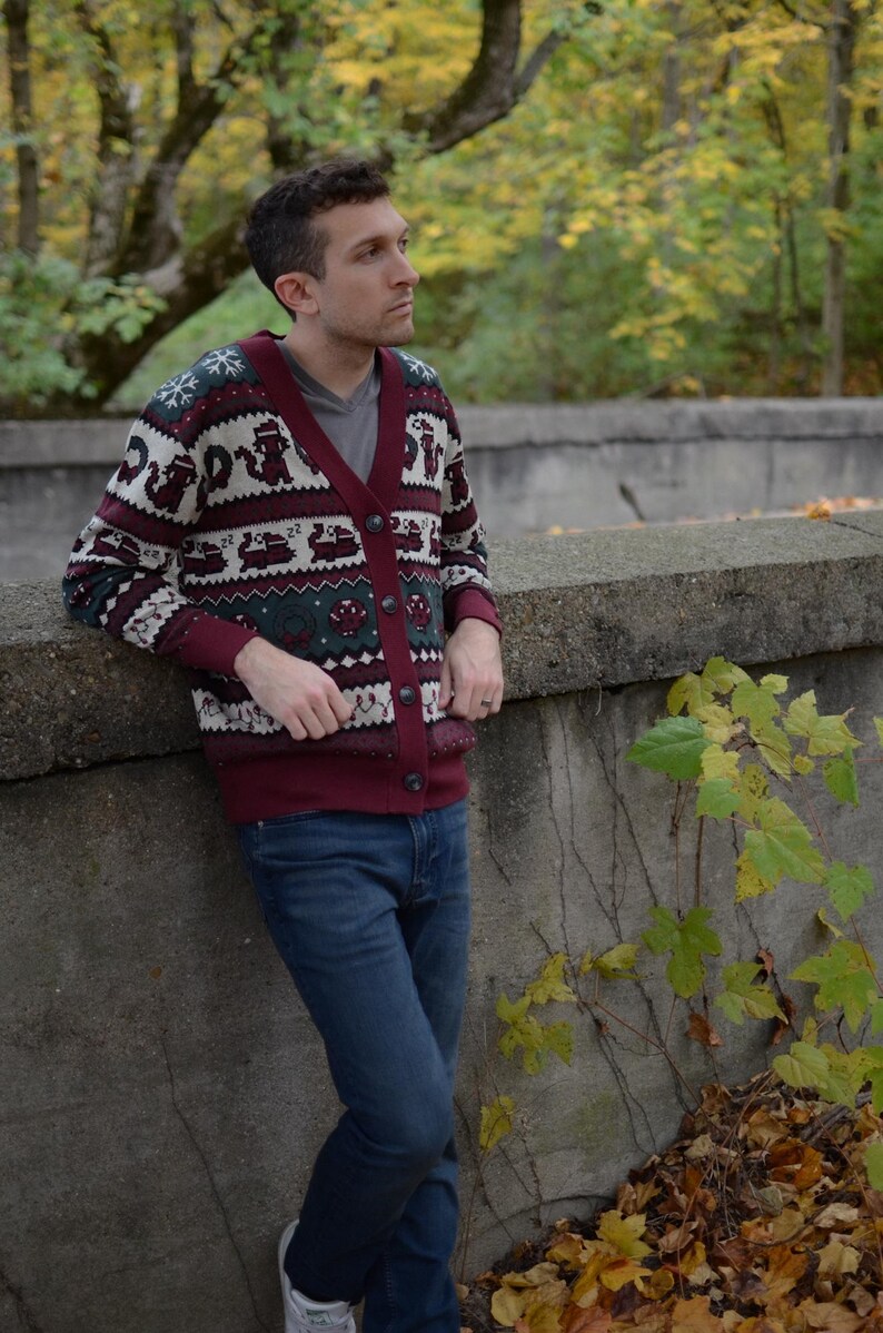 May include: A burgundy cardigan sweater with a festive pattern of reindeer, snowflakes, and other holiday motifs. The sweater has a button-up front and is paired with blue jeans. The background features a natural outdoor setting with trees and foliage.