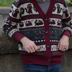 May include: A burgundy cardigan sweater with a festive pattern. The sweater features a V-neck, button closure, and a repeating design of pixelated holiday-themed images in white, green, and burgundy. The cuffs and trim are a solid burgundy color.