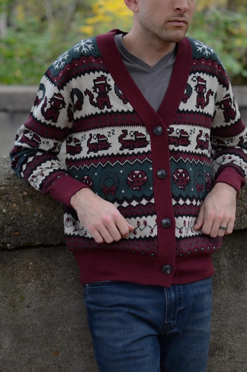 May include: A burgundy cardigan sweater with a festive pattern. The sweater features a V-neck, button closure, and a repeating design of pixelated holiday-themed images in white, green, and burgundy. The cuffs and trim are a solid burgundy color.