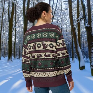 May include: A burgundy sweater with a festive pattern featuring snowflakes, wreaths, and train designs in green, cream, and burgundy. The sweater is worn by a person in a snowy forest setting.
