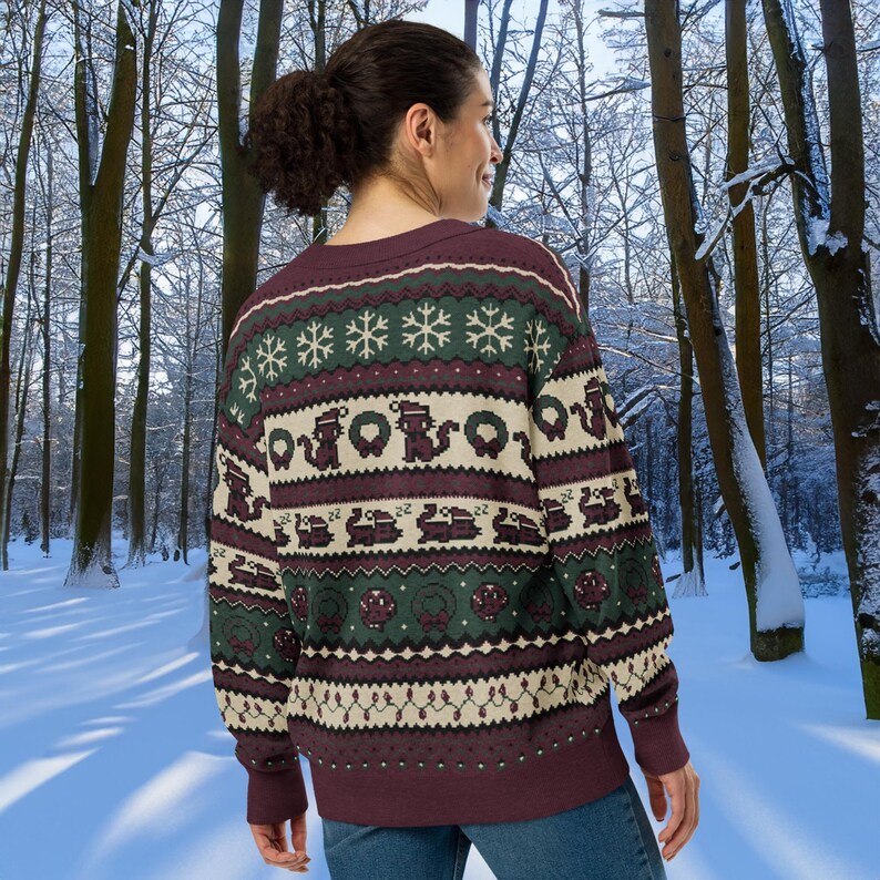 May include: A burgundy sweater with a festive pattern featuring snowflakes, wreaths, and train designs in green, cream, and burgundy. The sweater is worn by a person in a snowy forest setting.