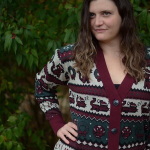 May include: A burgundy and cream-colored cardigan sweater with a festive pattern. The sweater features a V-neck, button closure, and long sleeves with a patterned design. The pattern includes snowflakes, reindeer, and other holiday-themed elements.