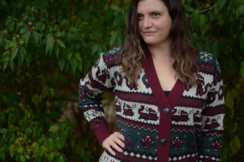 May include: A burgundy and cream-colored cardigan sweater with a festive pattern. The sweater features a V-neck, button closure, and long sleeves with a patterned design. The pattern includes snowflakes, reindeer, and other holiday-themed elements.