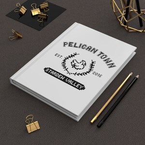May include: White notebook with a black and white logo that says "Pelican Town Est. 2015 Stardew Valley".