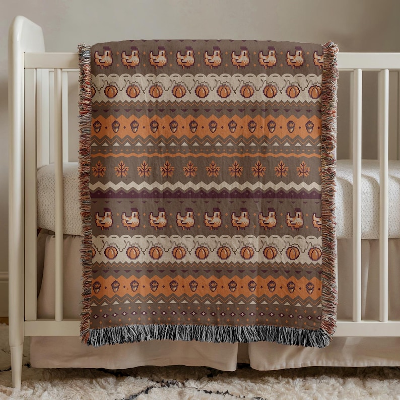 May include: A brown and orange woven throw blanket with a repeating pattern of turkeys, pumpkins, acorns, and leaves. The blanket is draped over a white crib, with a fringed edge.