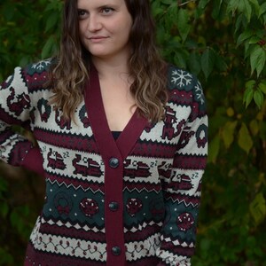 May include: Burgundy cardigan sweater with a festive pattern. The sweater features a deep V-neck, button closure, and a repeating design of snowflakes, wreaths, and other holiday-themed elements in green, white, and burgundy.