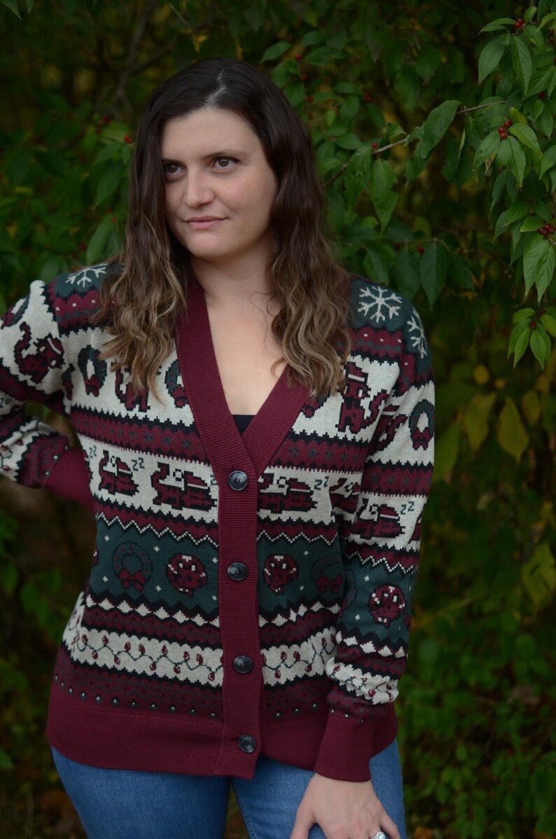 May include: Burgundy cardigan sweater with a festive pattern. The sweater features a deep V-neck, button closure, and a repeating design of snowflakes, wreaths, and other holiday-themed elements in green, white, and burgundy.